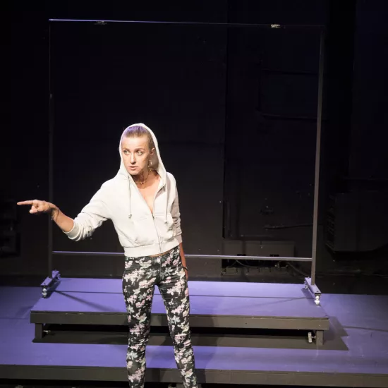 Zdjęcie przedstawia pierwszą próbę do spektaklu „Ifigenia ze Splott”. Przed podestami stoi Agnieszka Skrzypczak. Biodro aktorki jest lekko skręcone do boku, jej wzrok jest skierowany do boku. W tę samą stronę skierowany jest palec wskazujący prawej, zgiętej w łokciu ręki aktorki. Usta Agnieszki Skrzypczak są lekko otwarte a żuchwa wyciągnięta. Aktorka ma założony kaptur na głowę a za nią znajduje się scenografia w postaci trzepaka osiedlowego.