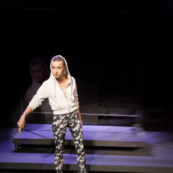 Zdjęcie przedstawia pierwszą próbę do spektaklu „Ifigenia ze Splott”. ”. Przed podestami stoi Agnieszka Skrzypczak. Biodro aktorki jest lekko skręcone do boku a wzrok jest skierowany w obiektyw. Palec wskazujący prawej ręki aktorki skierowany jest do dołu a usta są delikatnie otwarte. Aktorka ma założony kaptur na głowę a za nią znajduje się scenografia w postaci trzepaka osiedlowego.