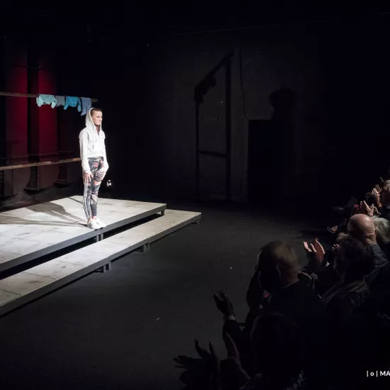 Zdjęcie przedstawia premierę spektaklu „Ifigenia ze Splott”. Agnieszka Skrzypczak znajduje się po lewej stronie fotografii. Jest odwrócona bokiem do obiektywu. Stoi wyprostowana, patrzy się na widownię przed sobą. Ma założony kaptur białej bluzy na głowie. Stoi na podeście, za nią widać scenografię trzepaka. Na górnej poprzeczce trzepaka wisi błękitna odzież. Aktorka i scena są oświetlone światłem reflektora. Po prawej stronie zdjęcia siedzi widownia, która patrzy na aktorkę i oklaskuje ją. 