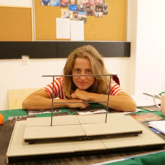 Zdjęcie przedstawia próbę stolikową do spektaklu „Ifigenia ze Splott”. Przy stole siedzi Agnieszka Skrzypczak z zgiętymi i rozłożonymi na stole łokciami. Głowa aktorki spoczywa na jej dłoniach blisko stołu a oczy skierowane są prosto w obiektyw. Przed aktorką na stole znajdują się fotografie i makieta osiedlowego trzepaka. Oprócz makiety po bokach, na stole znajduje się zeszyt, okulary, telefon, kubek, nożyczki, scenariusz i piórnik. Z tyłu wisi na ścianie tablica korkowa ze zdjęciami.  