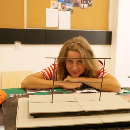 Zdjęcie przedstawia próbę stolikową do spektaklu „Ifigenia ze Splott”. Przy stole siedzi Agnieszka Skrzypczak z zgiętymi i rozłożonymi na stole łokciami. Głowa aktorki spoczywa na jej dłoniach blisko stołu a oczy skierowane są prosto w obiektyw. Przed aktorką na stole znajdują się fotografie i makieta osiedlowego trzepaka. Oprócz makiety po bokach, na stole znajduje się zeszyt, okulary, telefon, kubek, nożyczki, scenariusz i piórnik. Z tyłu wisi na ścianie tablica korkowa ze zdjęciami.  