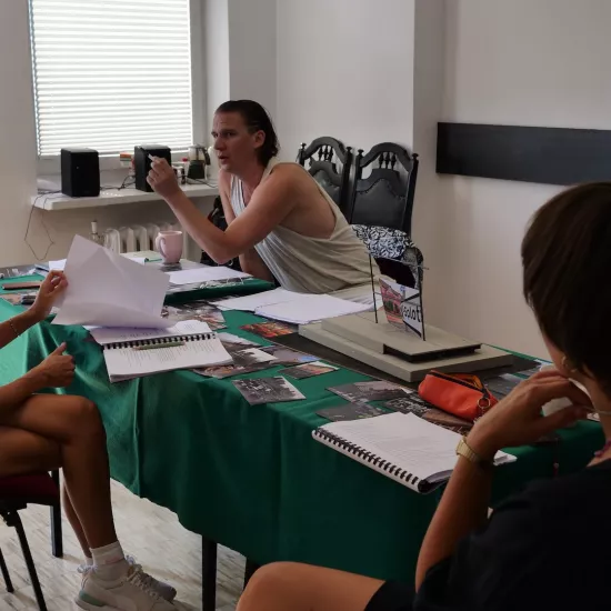 Zdjęcie przedstawia próbę stolikową do spektaklu „Ifigenia ze Splott”. Przy stole przodem siedzi reżyser Filip Gieldon a tyłem inspicjentka Marta Baraszkiewicz i Agnieszka Skrzypczak. Wzrok reżysera skierowany jest na aktorkę a jego lewa ręka jest zgięta w łokciu pod kątem prostym. Agnieszka Skrzypczak z lekko zgarbioną postawą przegląda scenariusz. Lewa ręka Marty Baraszkiewicz lekko dotyka jej brody. Na stole oprócz scenariuszy leży makieta trzepaka, fotografie, kubek i piórnik. 