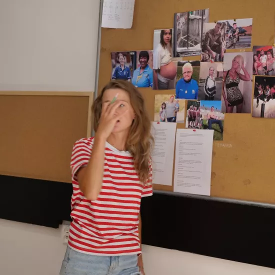 Zdjęcie przedstawia próbę stolikową do spektaklu „Ifigenia ze Splott”. Przy tablicy korkowej stoi Agnieszka Skrzypczak, której prawa ręka trzymająca długopis, zasłania jej prawie całą twarz. Na tablicy znajdują się fotografie różnych kobiet, mężczyzn i kota. Przypięta tam jest również mapa Cardiff oraz rożne teksty. 