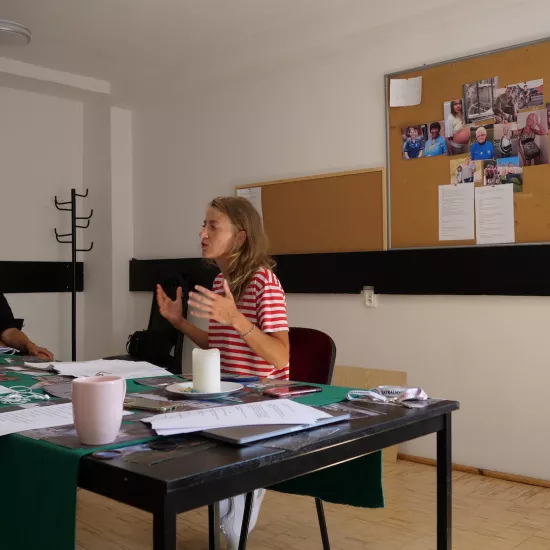 Zdjęcie przedstawia próbę stolikową do spektaklu „Ifigenia ze Splott”. Przy stole siedzi Agnieszka Skrzypczyk z inspicjentką Martą Baraszkiewicz. Aktorka trzyma swoje dłonie na wysokości klatki piersiowej nad stołem. Na samym stole znajduje się scenariusz, kubki, klucze, zdjęcia, świeczka, słuchawki i telefony. Z tyłu na ścianie wisi tablica korkowa ze zdjęciami różnych osób. 