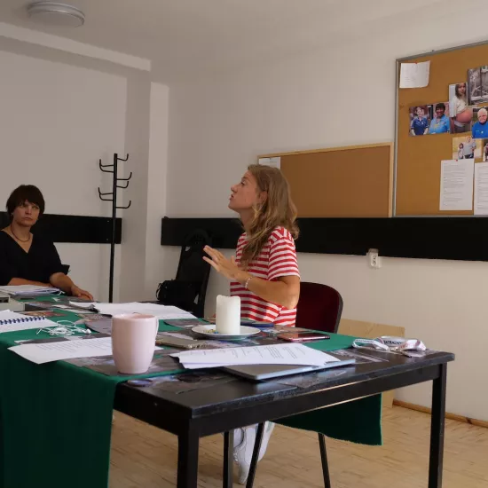 Zdjęcie przedstawia próbę stolikową do spektaklu „Ifigenia ze Splott”. Przy stole siedzi Agnieszka Skrzypczyk z inspicjentką Martą Baraszkiewicz. Głowa aktorki jest pochylona lekko do tyłu. Wzrok Marty Baraszkiewicz jest skierowany na Agnieszce Skrzypczyk. Na samym stole znajduje się scenariusz, kubki, klucze, zdjęcia, świeczka, słuchawki i telefony. Z tyłu na ścianie wisi tablica korkowa ze zdjęciami różnych osób. 