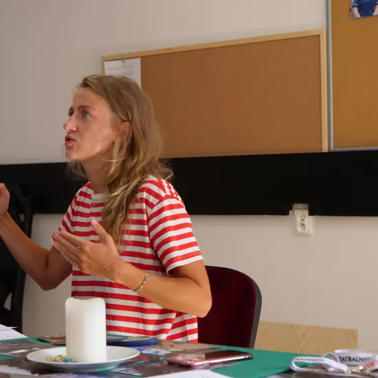 Zdjęcie przedstawia próbę stolikową do spektaklu „Ifigenia ze Splott”. Przy stole siedzi Agnieszka Skrzypczak ze zgiętymi, ale lekko wysuniętymi przed siebie rękami. Wzrok aktorki jest skierowany do boku, ponad obiektyw. Jej żuchwa jest wyciągnięta lekko do przodu a usta delikatnie otwarte. Przed aktorką na stole znajdują się fotografie, scenariusz, świeca, kubek i telefon. Z tyłu wisi na ścianie tablica korkowa ze zdjęciami.  