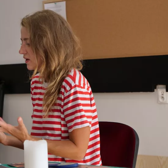 Zdjęcie przedstawia próbę stolikową do spektaklu „Ifigenia ze Splott”. Przy stole z wyciągniętymi przed siebie dłońmi siedzi Agnieszka Skrzypczyk. Wzrok aktorki skierowany jest w dół a jej usta są lekko otwarte. Na stole leży scenariusz a w tle widać tablice korkowe. 