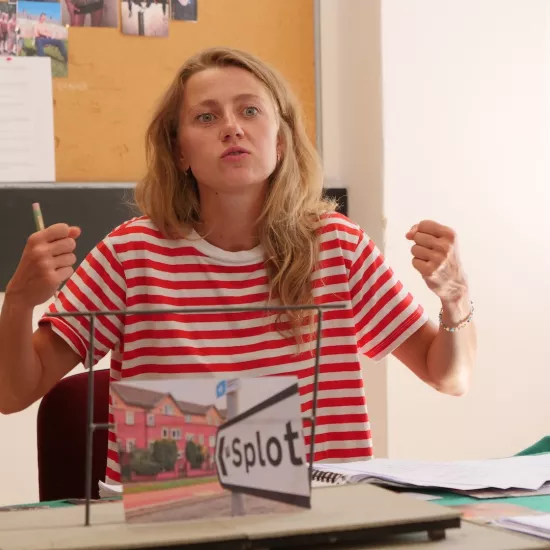 Zdjęcie przedstawia próbę stolikową do spektaklu „Ifigenia ze Splott”. Przy stole siedzi Agnieszka Skrzypczak z zgiętymi w łokciach i podniesionymi rękoma, których dłonie są zaciśnięte w pięści. Usta Agnieszki Skrzypczak są lekko ściągnięte i delikatnie otwarte. Przed aktorką na stole znajdują się fotografie i makieta osiedlowego trzepaka. Oprócz makiety na stole znajduje się zeszyt, okulary, scenariusz i nożyczki. 