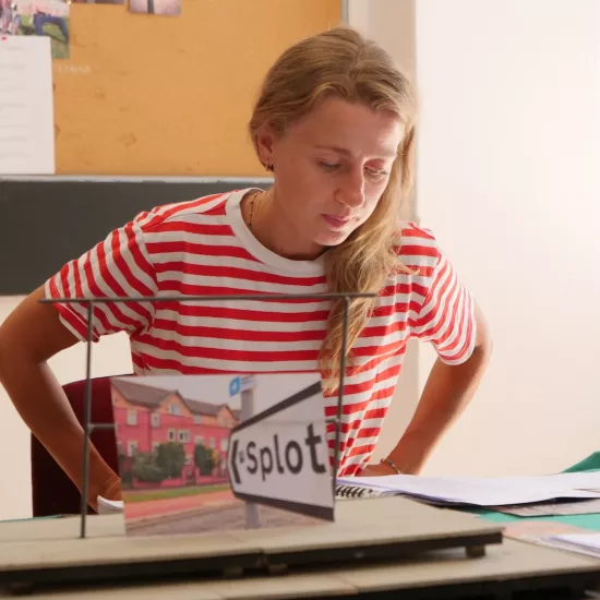 Zdjęcie przedstawia próbę stolikową do spektaklu „Ifigenia ze Splott”. Przy stole siedzi Agnieszka Skrzypczak z rękoma na biodrach. Wzrok aktorki utkwiony jest w scenariuszu a usta delikatnie ściągnięte do środka. Przed aktorką na stole znajdują się fotografie i makieta osiedlowego trzepaka. Oprócz makiety na stole znajduje się zeszyt, okulary, scenariusz i nożyczki. 