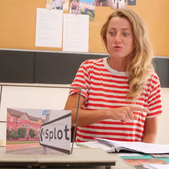 Zdjęcie przedstawia próbę stolikową do spektaklu „Ifigenia ze Splott”. Przy stole siedzi Agnieszka Skrzypczak. Wzrok aktorki utkwiony jest w makiecie trzepaka a jej dolna warga delikatnie wysunięta do przodu. Prawa ręka aktorki jest zgięta w łokciu i skierowana w bok, a wskazujący palec tej ręki jest wyprostowany. Przed aktorką na stole znajdują się fotografie i makieta osiedlowego trzepaka. 