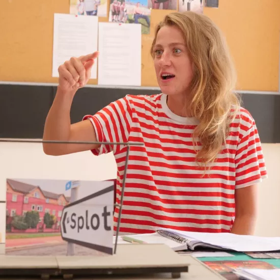 Zdjęcie przedstawia próbę stolikową do spektaklu „Ifigenia ze Splott”. Przy stole siedzi z lekko otwartymi ustami Agnieszka Skrzypczak. Wzrok aktorki utkwiony jest na wyprostowany palec wskazujący prawej, zgiętej w łokciu ręki. Przed aktorką na stole znajdują się fotografie i makieta osiedlowego trzepaka. 