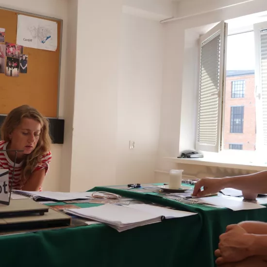 Zdjęcie przedstawia próbę stolikową do spektaklu „Ifigenia ze Splott”. Przy stole siedzi Agnieszka Skrzypczak i reżyser Filip Gieldon. Aktorka ze zgarbioną postawą patrzy w scenariusz przed sobą. Reżyser siedzi naprzeciwko aktorki, kierując swój wzrok na nią. Jego lewa ręka spoczywa na skrzyżowanych nogach a prawa na stole dociskając długopis. Na stole znajduje się makieta tapczanu ze zdjęciem angielskiej kamienicy i znakiem „Splott”, świeczka, okulary, nożyczki i scenariusze. Z tyłu wisi na ścianie tablica