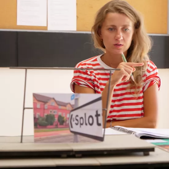 Zdjęcie przedstawia próbę stolikową do spektaklu „Ifigenia ze Splott”. Przy stole siedzi z lekko otwartymi ustami Agnieszka Skrzypczak. Wzrok aktorki utkwiony jest w przestrzeń poniżej linii obiektywu. W prawej, zgiętej w łokciu ręce trzyma długopis a palec wskazujący tej samej ręki jest wyprostowany. Przed aktorką na stole znajdują się fotografie, makieta osiedlowego trzepaka i scenariusz. 