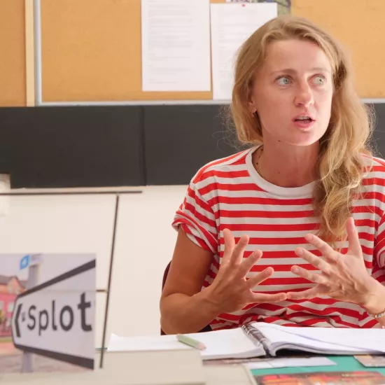 Zdjęcie przedstawia próbę stolikową do spektaklu „Ifigenia ze Splott”. Przy stole siedzi z lekko otwartymi ustami Agnieszka Skrzypczak. Wzrok aktorki utkwiony jest w przestrzeń po bocznej stronie obiektywu. Ręce Agnieszki Skrzypczak są zgięte w łokciach a palce dłoni mocno oddalone od siebie. Dłonie są zwrócone wewnętrzną częścią do aktorki. Przed aktorką na stole znajdują się fotografie, makieta osiedlowego trzepaka, okulary i scenariusz. Z tyłu widnieje tablica korkowa z tekstami i zdjęciami.