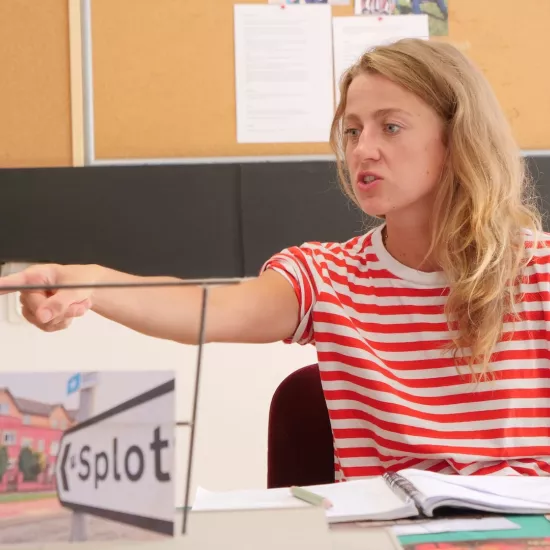 Zdjęcie przedstawia próbę stolikową do spektaklu „Ifigenia ze Splott”. Przy stole siedzi z lekko otwartymi ustami i zaciśniętymi zębami Agnieszka Skrzypczak. Wzrok aktorki utkwiony jest w przestrzeń poniżej linii obiektywu. Prawa ręka aktorki jest wyprostowana a palec wskazujący jest wyciągnięty do przodu. Przed aktorką na stole znajdują się fotografie, makieta osiedlowego trzepaka, okulary i scenariusz. Z tyłu widnieje tablica korkowa z tekstami i zdjęciami.