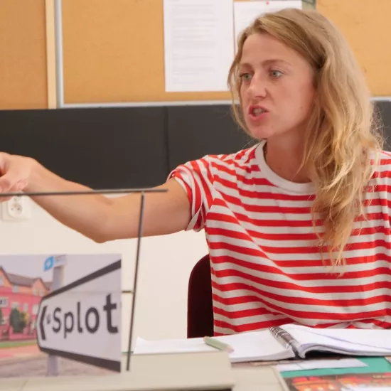 Zdjęcie przedstawia próbę stolikową do spektaklu „Ifigenia ze Splott”. Przy stole siedzi z lekko otwartymi ustami i zaciśniętymi zębami Agnieszka Skrzypczak. Wzrok aktorki utkwiony jest w przestrzeń poniżej linii obiektywu. Prawa ręka aktorki jest wyprostowana a palec wskazujący jest wyciągnięty do przodu. Przed aktorką na stole znajdują się fotografie, makieta osiedlowego trzepaka, okulary i scenariusz. Z tyłu widnieje tablica korkowa z tekstami i zdjęciami.