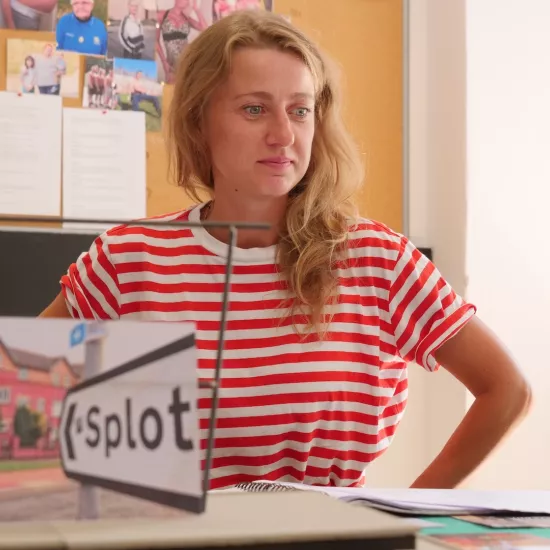 Zdjęcie przedstawia próbę stolikową do spektaklu „Ifigenia ze Splott”. Przy stole siedzi Agnieszka Skrzypczak z założonymi na biodra rękoma. Wzrok aktorki skierowany jest w przestrzeń, poniżej linii obiektywu. Przed Agnieszką Skrzypczak na stole znajdują się fotografie i makieta osiedlowego trzepaka z opartym na niej zdjęciem brytyjskiej kamienicy. Z tyłu na ścianie wisi tablica korkowa z różnymi zdjęciami i tekstami. 