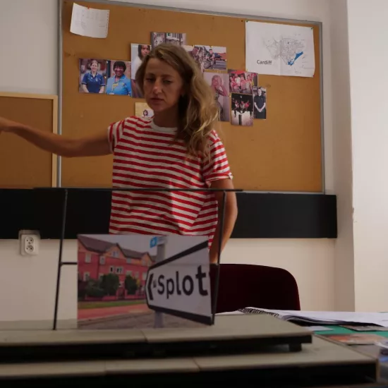 Zdjęcie przedstawia próbę stolikową do spektaklu „Ifigenia ze Splott”. Przed stołem z podniesioną prawą ręką i wyprostowanym palcem wskazującym stoi Agnieszka Skrzypczak. Natomiast lewa ręka aktorki spoczywa wzdłuż ciała. Wzrok aktorki jest spuszczony w dół, tak samo kącki ust. Na stole znajdują się fotografie i makieta osiedlowego trzepaka z opartym na niej zdjęciem brytyjskiej kamienicy. Z tyłu na ścianie wisi tablica korkowa z różnymi zdjęciami i tekstami. 