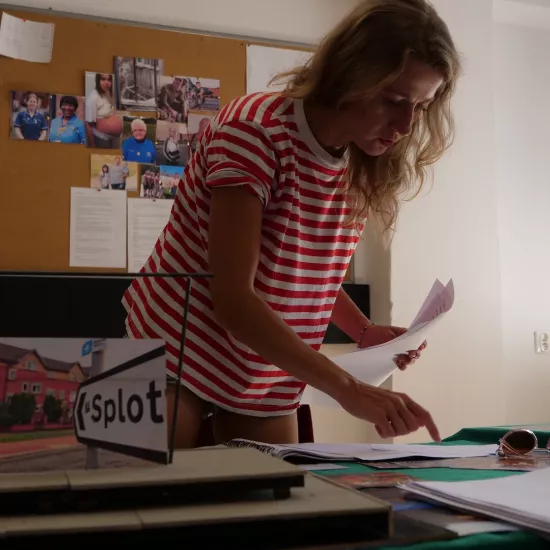 Zdjęcie przedstawia próbę stolikową do spektaklu „Ifigenia ze Splott”. Nad stołem pochyla się Agnieszka Skrzypczak. W lewej ręce trzyma kartki papieru, natomiast palcem wskazującym prawej ręki dotyka innych leżących kartek. Wzrok aktorki utkwiony jest na kartkach. Na stole znajdują się fotografie, okulary, scenariusz i makieta osiedlowego trzepaka z opartym na niej zdjęciem brytyjskiej kamienicy. Z tyłu na ścianie wisi tablica korkowa z różnymi zdjęciami i tekstami. 