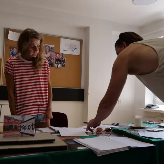 Zdjęcie przedstawia próbę stolikową do spektaklu „Ifigenia ze Splott”. Przy stole stoi reżyser Filip Gieldon wraz z Agnieszką Skrzypczak. Reżyser nachyla się nad stołem, dotykając palcem lewej ręki zdjęcia na stole. W tej samej ręce trzyma czarny długopis. Wzrok aktorki utkwiony jest w miejscu wskazywanym przez reżysera a jej ręce spoczywają wzdłuż ciała. Na stole znajdują się fotografie, okulary, świeca, kubek, scenariusz i makieta osiedlowego trzepaka z opartym na niej zdjęciem brytyjskiej kamienicy. 