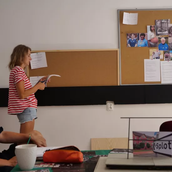 Zdjęcie przedstawia próbę stolikową do spektaklu „Ifigenia ze Splott”. Agnieszka Skrzypczak stojąc bocznym profilem tnie nożyczkami kartkę. Po lewej stronie aktorki wiszą na ścianie dwie tablice korkowe z tekstami i zdjęciami różnych osób. Przy stole, również bocznym profilem siedzi ubrana na ciemno, z ręką na kolanie Marta Baraszkiewicz. Obie kobiety znajdują się po lewej stronie fotografii. Na jej środku mieści się stół, na którym leży kubek, piórnik, fotografie, makieta trzepaka z opartym na nim zdjęciem