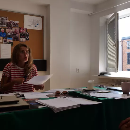 Zdjęcie przedstawia próbę stolikową do spektaklu „Ifigenia ze Splott”. Przy stole siedzi reżyser Filip Gieldon wraz z Agnieszką Skrzypczak. Reżyser siedzi z rękoma położonymi na swoich kolanach. Aktorka siedząc naprzeciwko reżysera trzyma w prawej ręce nożyczki a w lewej kartkę papieru. Wzrok Agnieszki Skrzypczak skierowany jest na reżyser. Jej oczy są mocno wytrzeszczone a usta ściągnięte i lekko otwarte.  Na stole znajdują się fotografie, okulary, świeca, kubek, scenariusz i makieta osiedlowego trzepaka.