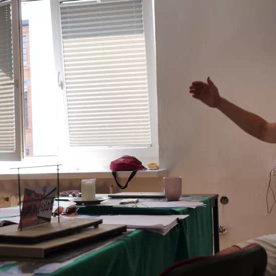 Zdjęcie przedstawia próbę stolikową do spektaklu „Ifigenia ze Splott”. Przy stole siedzi reżyser Filip Gieldon a naprzeciwko niego z rękami wzdłuż ciała stoi Agnieszka Skrzypczak. Reżyser podnosząc prawą rękę do góry utrzymuje kontakt wzrokowy z aktorką. Usta reżysera są otwarte a głowa pochylona lekko do tyłu. W swojej lewej ręce trzyma czarny długopis. Na stole znajdują się fotografie, okulary, świeca, kubek, scenariusz i makieta osiedlowego trzepaka z opartym na niej zdjęciem brytyjskiej kamienicy.