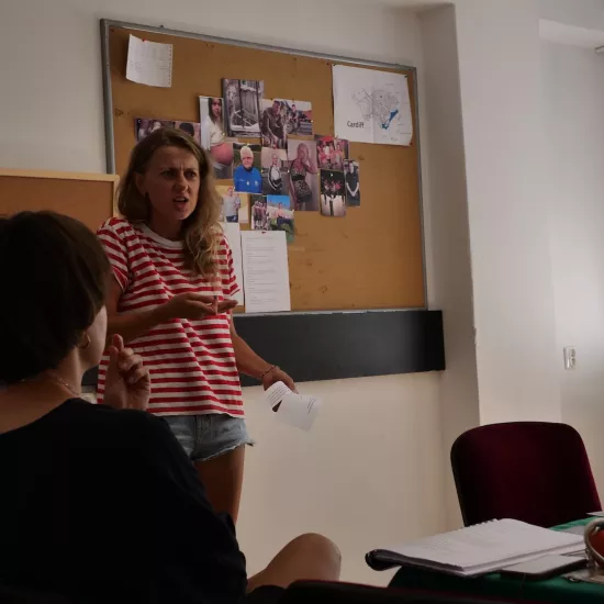 Zdjęcie przedstawia próbę stolikową do spektaklu „Ifigenia ze Splott”. Przy stole, tyłem do obiektywu siedzi inspicjentka Marta Baraszkiewicz a obok niej stoi Agnieszka Skrzypczak. Aktorka w lewej ręce trzyma tekst a prawą ma podniesioną na wysokość klatki. Jej brwi są zmarszczone, usta otwarte, ramiona podniesione do góry a wzrok skierowany przed siebie. Lewa ręka inspicjentki jest podniesiona na wysokość brody. Na stole znajduje się scenariusz, kubek, telefon i piórnik. Z tyłu wisi tablica krokowa. 