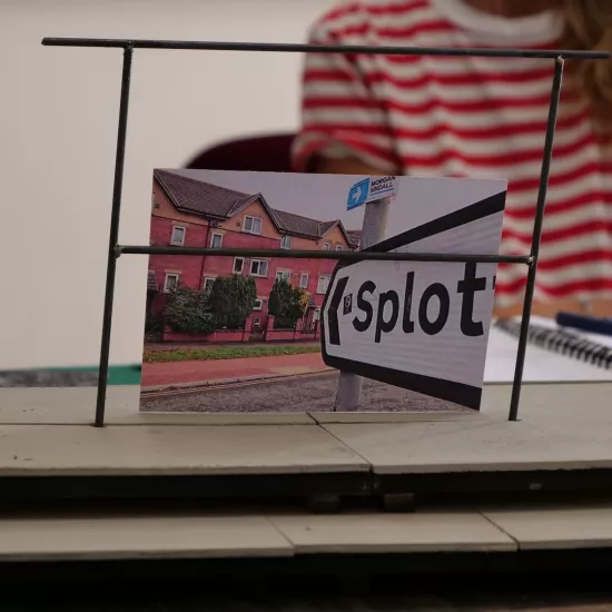 Zdjęcie przedstawia próbę stolikową do spektaklu „Ifigenia ze Splott”. Zdjęcie skoncentrowane na makiecie osiedlowego trzepaka, na którym opiera się fotografia brytyjskiej kamienicy, którą poprzedza znak z nazwą dzielnicy Splott. W tle został uchwycony tułów aktorki z jej koszulką w czerwono- białe paski. Na drugim planie można, również dostrzec scenariusz, nożyczki i fotografie.  