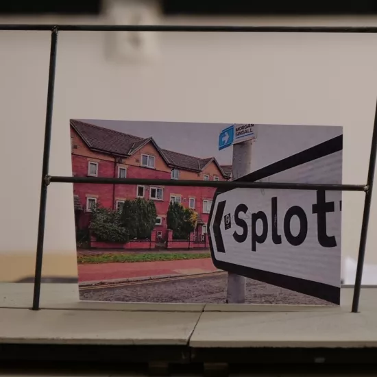 Zdjęcie przedstawia próbę stolikową do spektaklu „Ifigenia ze Splott”. Zdjęcie skoncentrowane na makiecie osiedlowego trzepaka, na którym opiera się fotografia brytyjskiej kamienicy, którą poprzedza znak z nazwą dzielnicy Splott. W tle został uchwycony tułów aktorki z jej koszulką w czerwono- białe paski. Na drugim planie można, również dostrzec scenariusz, nożyczki i fotografie.  