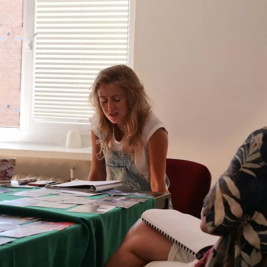 Zdjęcie przedstawia próbę stolikową do spektaklu „Ifigenia ze Splott”. Przy stole siedzi przodem Agnieszka Skrzypczak i tyłem reżyser Filip Gieldon. Na stole przed aktorką leży otwarty scenariusz i fotografie. Oczy aktorki są zamknięte a  ręce wyprostowane w łokciach, oparte o siedzenie. Reżyser trzyma na kolanach scenariusz a jego wzrok skierowany jest przed siebie. Na stole znajdują się także kubki, okulary i telefon. 