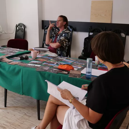 Zdjęcie przedstawia próbę stolikową do spektaklu „Ifigenia ze Splott”. ”. Przy stole siedzi od lewej strony trzymająca ręce na biodrach Agnieszka Skrzypczak, po środku z palcem wskazującym skierowanym ku górze reżyser Filip Gieldon, naprzeciwko aktorki scenografka i kostiumografka Adrianna Gołębiewska  i po prawej trzymająca scenariusz na nogach inspicjentka Marta Baraszkiewicz. Na stole leży dużo fotografii, scenariusz, piórnik, kubek i okulary. Przy stole leżą buty Agnieszki Skrzypczak i butelka soku.