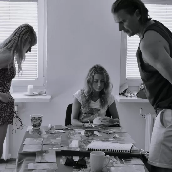 Zdjęcie przedstawia próbę stolikową do spektaklu „Ifigenia ze Splott”. Zdjęcie jest czarno-białe.  Przy stole siedzi Agnieszka Skrzypczak, która przegląda scenariusz. Nad tym samym stołem pochyla się reżyser Filip Gieldon wraz z scenografką Adrianną Gołębiewską. Spoglądają oni na rozłożone na stole zdjęcia. Oboje są skierowani bokiem do obiektywu, po środku nich przodem siedzi Agnieszka Skrzypczak. Na stole oprócz zdjęć znajdują się kubki, okulary i telefon. 