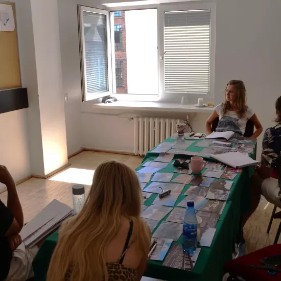 Zdjęcie przedstawia próbę stolikową do spektaklu „Ifigenia ze Splott”. Fotografia obejmuje dużą część pomieszczenia w tym cały stół, przy którym siedzi reżyser Filip Gieldon, inspicjentka Marta Baraszkiewicz, Agnieszka Skrzypczak i scenografka Adrianna Gołębiewska. Aktorka, która spogląda na tablicę korkową jako jedyna jest przodem do obiektywu, natomiast pozostałe osoby są zwrócone tyłem. Na stole leży dużo fotografii, kubki, butelki i scenariusze