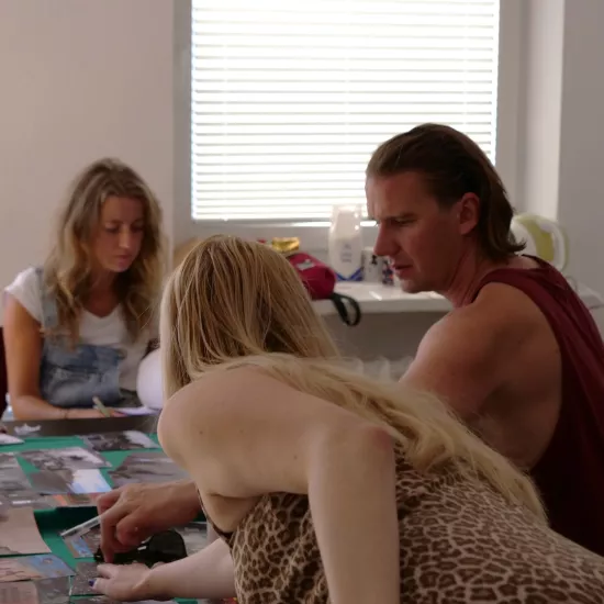 Zdjęcie przedstawia próbę stolikową do spektaklu „Ifigenia ze Splott”. Przy stole siedzi Agnieszka Skrzypczak oraz reżyser Filip Gieldon. Nad stołem, tyłem do obiektywu pochyla się scenografka Adrianna Gołębiewska, która wraz z reżyserem układają zdjęcia na stole.