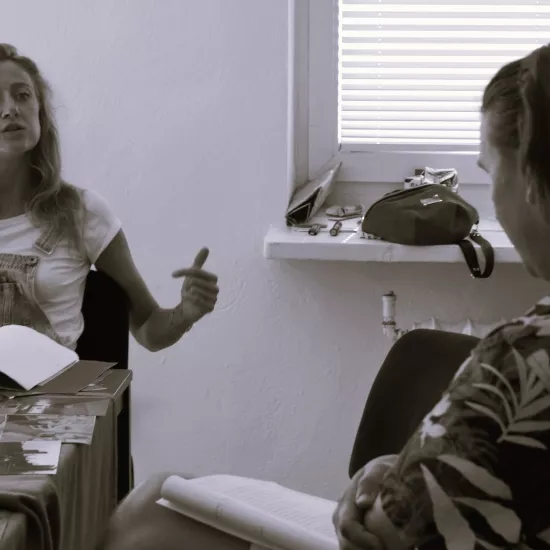 Zdjęcie przedstawia próbę stolikową do spektaklu „Ifigenia ze Splott”. Zdjęcie jest czarno- białe. Przy stole siedzi przodem Agnieszka Skrzypczak i tyłem reżyser Filip Gieldon. Na stole przed aktorką leży otwarty scenariusz i fotografie. Usta aktorki są lekko otwarte i głowa lekko pochylona do tyłu. Lewa ręka jest lekko wyciągnięta do boku a palec wskazujący i kciuk tej ręki wyprostowany. Kontakt wzrokowy skierowany jest na reżysera, który na swoich kolanach trzyma scenariusz. 