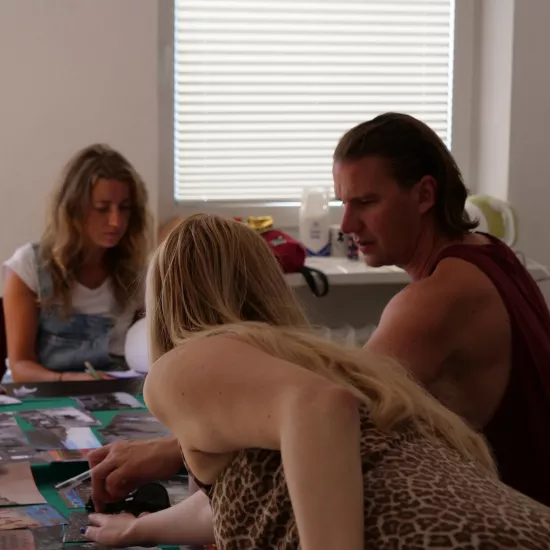 Zdjęcie przedstawia próbę stolikową do spektaklu „Ifigenia ze Splott”. Przy stole siedzi Agnieszka Skrzypczak oraz reżyser Filip Gieldon. Nad stołem, tyłem do obiektywu pochyla się scenografka Adrianna Gołębiewska, która wraz z reżyserem układają zdjęcia na stole.