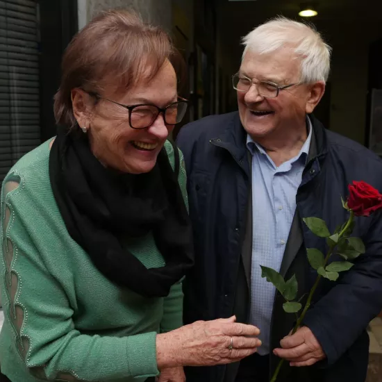Zdjęcie przedstawia dwójkę starszych uśmiechniętych ludzi - kobietę i mężczyznę. Mężczyzna trzyma w ręku czerwoną różę. 