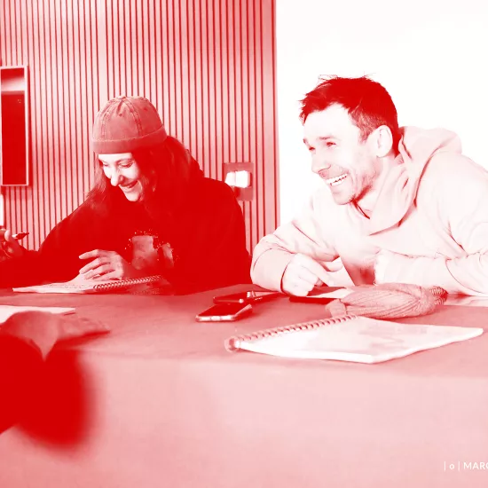 Zdjęcie przedstawia pierwszą próbę spektaklu „Morderstwo w hotelu". Po lewej stronie stołu siedzi aktor Mariusz Witkowski. Na przeciwko niego, po drugiej stronie stołu siedzą aktorzy Natalia Klepacka i Krzysztof Wach. Aktorka ma na głowie czapkę. Wszyscy aktorzy się śmieją. Na zdjęcie został nałożony czerwony filtr.