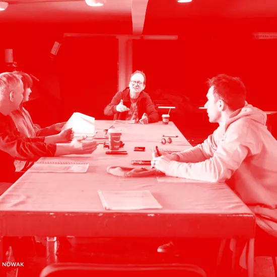 zdjęcie przedstawia pierwszą próbę spektaklu "Morderstwo w hotelu". Przy stole siedzą aktorzy, inspicjentka i reżyser. Po lewej stronie siedzą aktor Mariusz Witkowski i inspicjentka Marta Baraszkiewicz, po środku na szczycie stołu siedzi reżyser Jacek Bończyk, który coś tłumaczy. Po prawej stronie siedzą aktorzy Natalia Klepacka i Krzysztof Wach. Na stole znajdują się egzeplarze tekstu. Na zdjęcie nałożony został czerwony filtr.