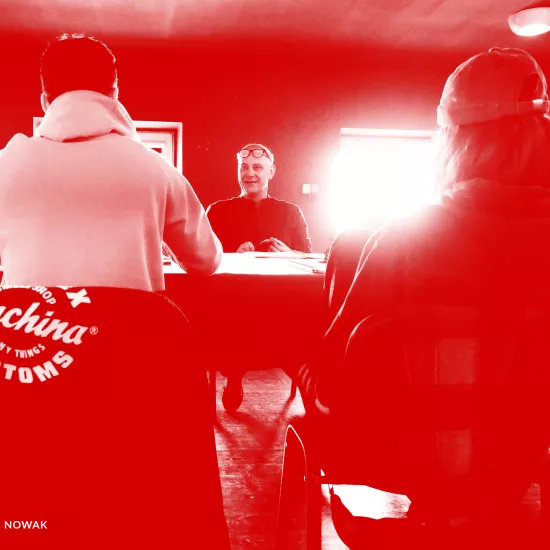 Zdjęcie przedstawia pierwszą próbę spektaklu „Morderstwo w hotelu". Tyłem do obiektywu siedzą przy stole aktorzy Krzysztof Wach i Natalia Klepacka (ona w czapce na głowie). Przodem do obiektywu, po drugiej stronie stołu siedzi aktor Mariusz Witkowski. Za nim znajdują się dwa okna, przez które przebija mocne światło słoneczne. Na zdjęcie został nałożony czerwony filtr.