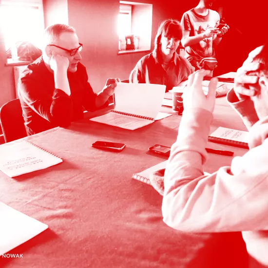 zdjęcie przedstawia pierwszą próbę spektaklu „Morderstwo w hotelu". Po lewej stronie stołu siedzi aktor Mariusz Witkowski czytający egzemplarz sztuki i przerzuca jego kartkę. Obok siedzi inspicjentka Marta Baraszkiewicz. W tle widać pracownika teatru Marcina Majczaka. Po prawej stronie blisko obiektywu siedzi aktor Krzystzof Wach zasłaniający prawą ręką swoją głowę. Lewą rekę ma uniesioną. Na zdjęcie został nałożony czerwony filtr. 