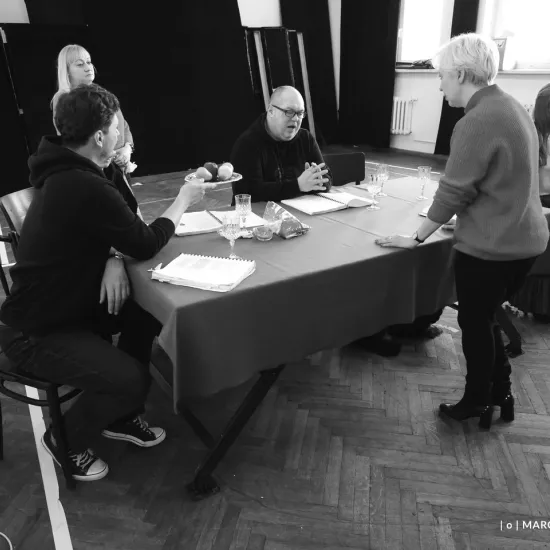 zdjęcie przedstawia próbę spektaklu „Świętoszek". Zostało zrobione na dużej sali prób w Teatrze Jaracza w Łodzi. Aktorzy Marcin Włodarski i Paweł Tucholski siedzą przy stole. Marcin Włodarski podaje Pawłowi Tucholskiemu tacę z owocami. Na stole są puste kieliszki do wina, które są rekwizytami w spektaklu oraz egzemplarze scenariusza. W tle widać stojącą inspicjentkę Katarzynę Brudek. Na krześle z boku przy stole siedzi aktorka Matylda Paszczenko. Nad stołem stoi reżyserka Magdalena Piekorz 