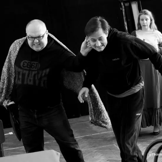 zdjęcie przedstawia próbę spektaklu „Świętoszek" w reżyserii Magdaleny Piekorz, na zdjęciu Paweł Tucholski prowadzi aktora Marka Nędzę ciągnąc go za ucho, w tle stoi aktorka Matylda Paszczenko. Autorem zdjęcia jest Marcin Nowak