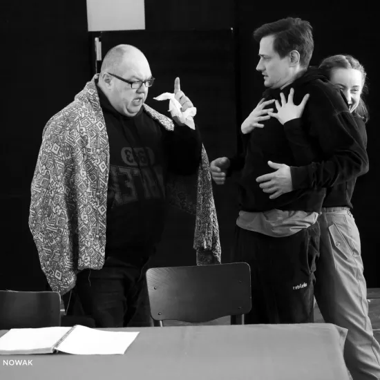 zdjęcie przedstawia próbę spektaklu „Świętoszek". Zostało zrobione na dużej sali prób w Teatrze Jaracza w Łodzi. Bokie do aparatu, za stołem stoi aktor Paweł Tucholski z uniesioną dłonią, w której trzyma chusteczkę. Na przeciwko niego stoi aktor Marek Nędza, za nim stoi aktorka Iwona Dróżdż, która go przytula od tyłu, trzymając dłonie na jego klatce piersiowej. 