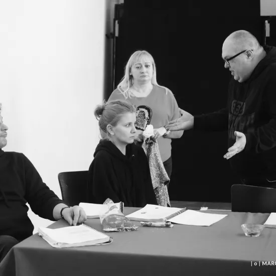 zdjęcie przedstawia próbę spektaklu „Świętoszek". Zostało zrobione na dużej sali prób w Teatrze Jaracza w Łodzi. Przy stole siedzą aktorzy Marcin Włodarski, Sara Lityńska. Nad nimi stoi aktor Paweł Tucholski. W tle stoi inspicjentka Katarzyna Brudek. 