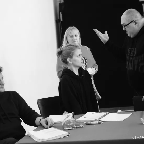 zdjęcie przedstawia próbę spektaklu „Świętoszek". Zostało zrobione na dużej sali prób w Teatrze Jaracza w Łodzi. Przy stole siedzi aktor Marcin Włodarski. Obok aktorka Sara Lityńska, nad nią stoi Paweł Tucholski z uniesioną prawą ręką jakby krzyczał na aktorkę. Za nimi stoi inspicjentka Katarzyna Brudek. 