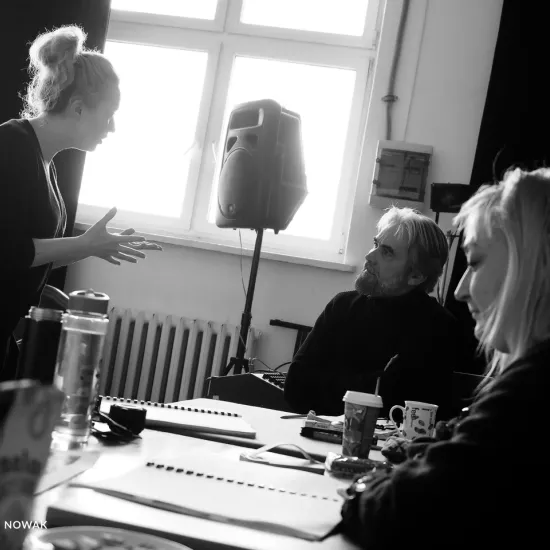 zdjęcie przedstawia próbę spektaklu „Świętoszek" w reżyserii Magdaleny Piekorz. Na zdjęciu na pierwszym planie, bokiem widać inspicjentkę Katarzynę Brudek, która siedzi za stołem. Obok siedzi aktor Dariusz Bereski. Przed nimi stoi aktorka Iwona Karlicka, która coś w tłumaczy. 