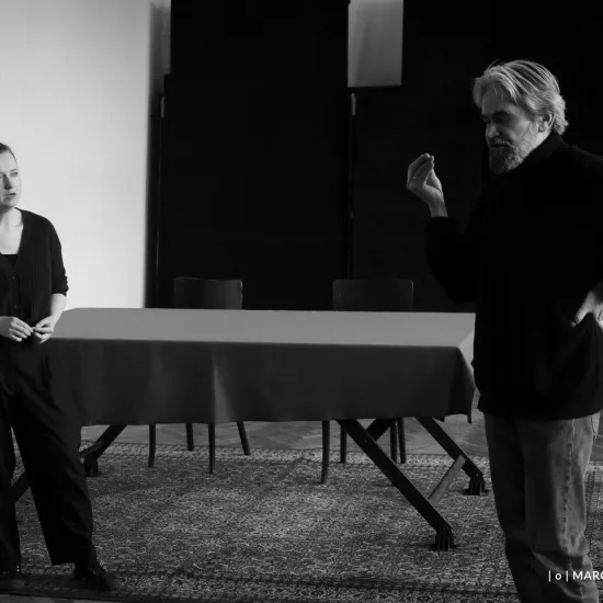 zdjęcie przedstawia próbę spektaklu „Świętoszek" w reżyserii Magdaleny Piekorz. Na zdjęciu jest dwójka aktorów: po lewej stronie Iwona Karlicka wcielająca się w rolę Doryny, ubrana na czarno, stoi w dużej odległośc od aktora Dariusza Bereskiego wcielającego się w rolę Orgona. Stoją przodem do siebie. Dariusz Bereski jest lewą ręką podparty pod bokiem, lewą ręką gestykuluje. Ma na sobie czarny swetr i spodnie dżinsowe. W tle widać stół nakryty suknem. Zdjęcie jest czarnobiałe.