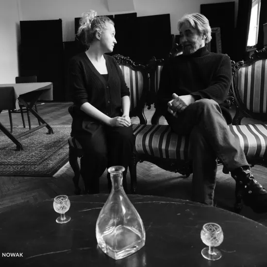 zdjęcie przedstawia próbę spektaklu „Świętoszek" w reżyserii Magdaleny Piekorz. Na zdjęciu dwoje aktorów: tyłem stoi Dariusz Bereski, z przodu widzimy Iwonę Karlicką. Przed nimi stoi niski stolik, na którym stoi pusta karafka i dwa kieliszki. 